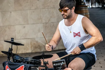 El batería, con camiseta blanca de tirantes y gafas de sol, toca una batería electrónica.
