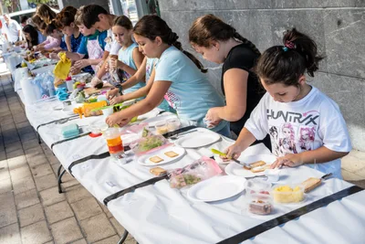 Una fila de chavales trabaja a lo largo de la mesa con mantel blanco, entre táperes y platos de papel.