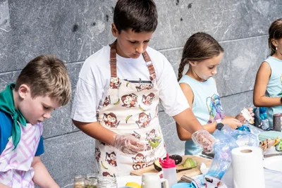 Un chico con delantal de monos y guantes de plástico prepara su pintxo; a su lado, una niña con trenzas.