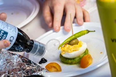 Unas manos terminan un pintxo de huevo relleno con guindilla y aceituna, con un chorro de crema balsámica.