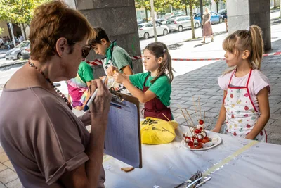 Una mujer del jurado con carpeta mira el plato de dos niñas con delantal, que esperan a su lado.