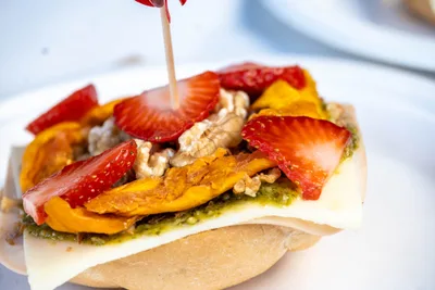 Un pintxo de queso con pimiento asado, nueces y fresas, coronado por un palillo con una fresa de papel.