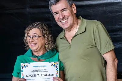 Una mujer con pañuelo verde enseña el diploma de accésit del concurso fotográfico junto a un hombre sonriente.