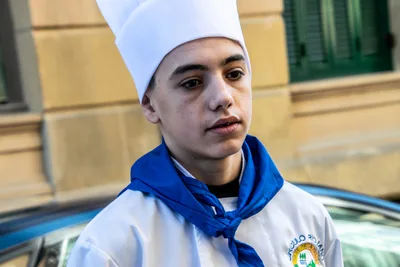 Retrato de un tamborrero joven con gorro de cocinero y pañuelo azul, delante de una fachada de piedra.