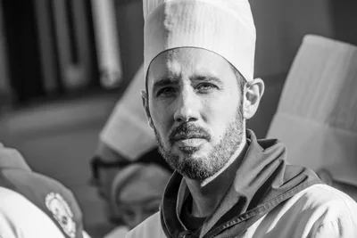 Retrato en blanco y negro de un cocinero con barba y gorro alto, entre otros tamborreros desenfocados.