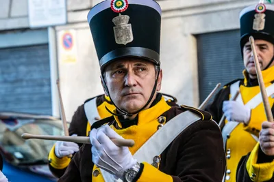 Un gastador mayor con chacó y guantes blancos toca el tambor con las baquetas en alto.