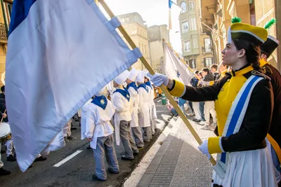 Una cantinera hace ondear la bandera blanca y azul mientras la compañía de cocineros baja la calle detrás.