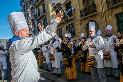 Un cocinero dirige con los dos brazos abiertos delante de la formación, en una calle con balcones.