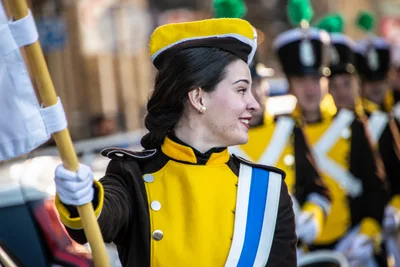 Una cantinera sostiene el asta de la bandera, con uniforme amarillo y marrón y banda azul y blanca.