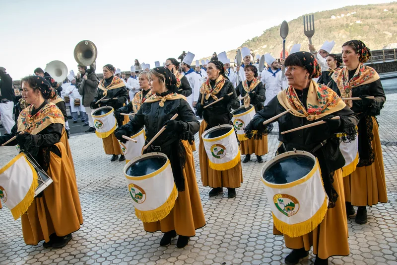La fila de mujeres con mantón y falda amarilla toca el barril junto al paseo, con el monte al fondo.