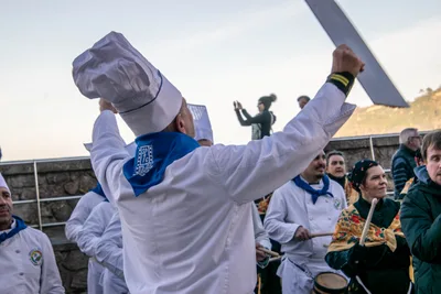 Un cocinero de espaldas levanta los brazos con una cuchilla de cartón, junto al pretil del paseo.