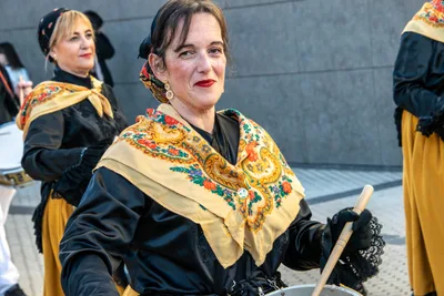 Una mujer con mantón y pendientes largos toca el barril, mirando al frente.