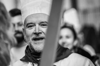 Retrato en blanco y negro de un cocinero con barba cantando, medio tapado por un asta.