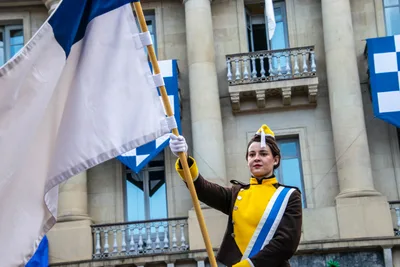 Una cantinera sostiene la bandera blanca y azul delante de una fachada con banderas colgadas en los balcones.