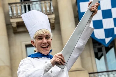 Una cocinera ríe levantando la cuchilla de cartón, con las banderas de la plaza al fondo.