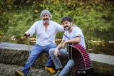 Dos hombres sonrientes sentados en un murete de piedra junto a un río con hojas de otoño en el agua. El de la izquierda, de pelo y barba canosos y camisa blanca, apoya las manos en un bastón de colores; el de la derecha, con camiseta blanca, sostiene una trikitixa de fuelle rojo.