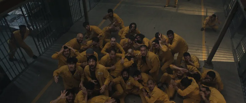«Ghost in the Cell» filmeko fotograma: goitik ikusitako kartzela-patio bat, non mono horiak jantzitako hamarnaka preso lurrean pilatzen diren, elkarri helduta, burdin-hesi batzuen ondoan.